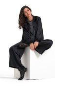 A woman with long dark hair sits on a white cube pedestal against a solid white studio background. She is wearing a black wide-sleeve satin-like top, matching black wide-leg trousers, and black ankle boots with silver studs.