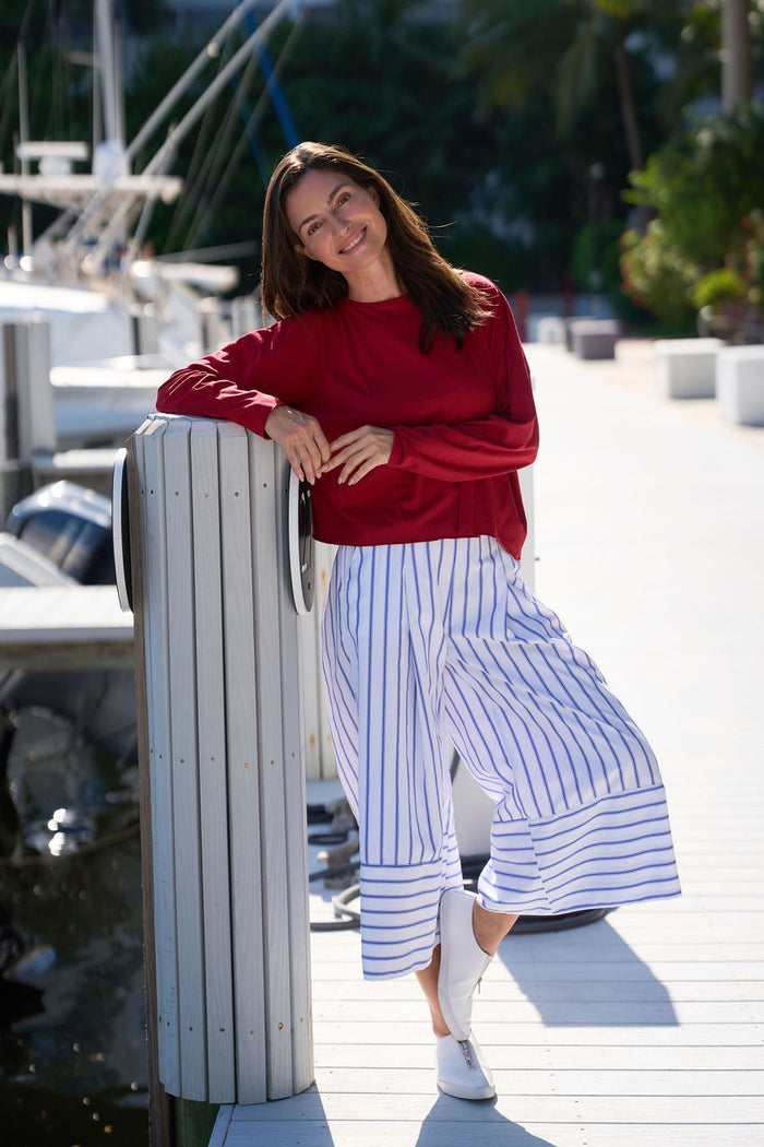 Woman in a red pima cotton shirt and striped pants standing by a marina with boats in the background.
