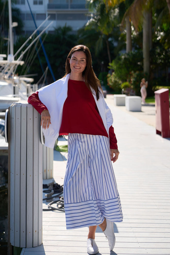 Woman in a red pima cotton shirt and white sweatshirt with a striped pants standing by a marina with boats and buildings in the background.