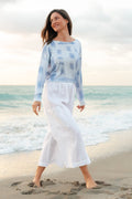 Woman in a light blue tie-dye top and white pants standing on a beach.