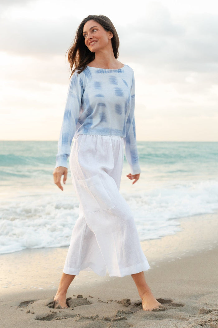 Woman in a light blue tie-dye top and white pants standing on a beach.