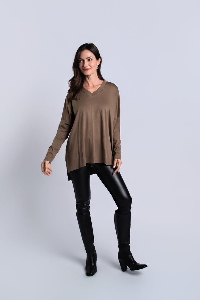 A woman modeling a taupe brown long-sleeve V-neck tunic top, paired with black faux-leather pants and black knee-high boots, against a light gray studio background.