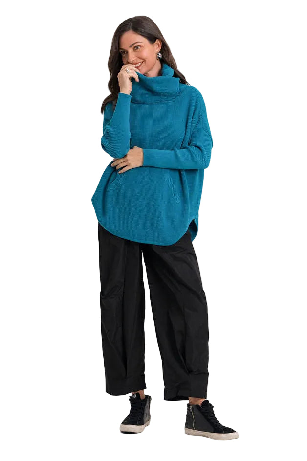 A woman modeling a teal blue pima cotton cowl-neck sweater, black wide-leg cropped pants, and black sneakers against a plain white studio background.
