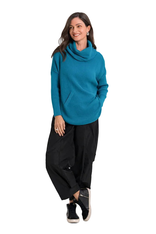 A woman modeling a teal blue cowl-neck knit pima cotton sweater and black wide-leg pants against a plain white studio background.