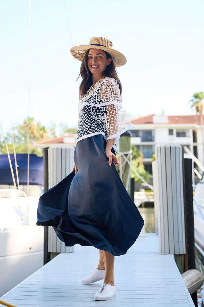 Woman in a white lace top and blue skirt standing on a dock with a sailboat in the background.