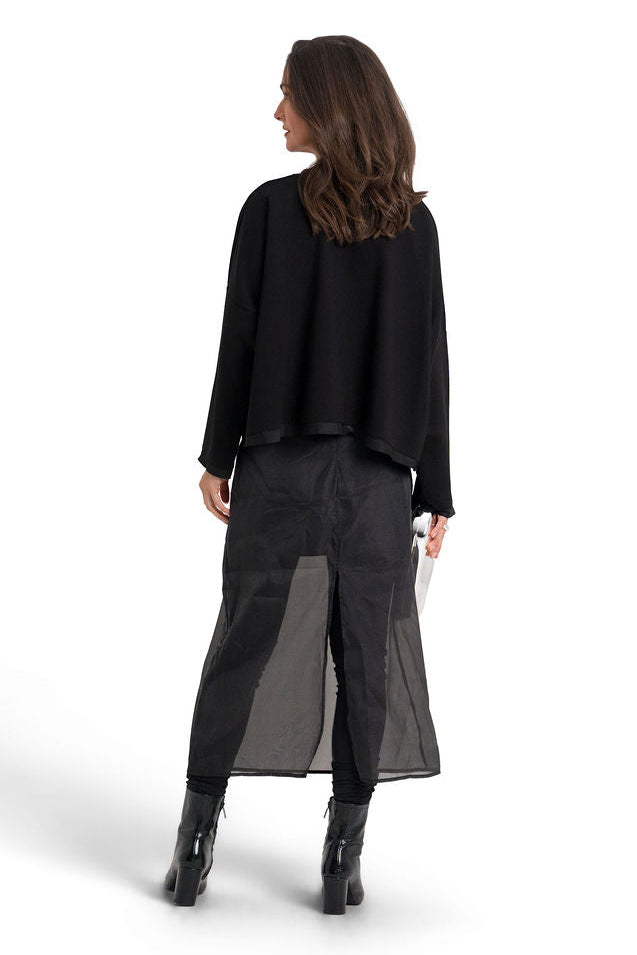 A woman shown from the back wearing a black long-sleeve top, a sheer black midi skirt, and black ankle boots, standing against a solid white background.
