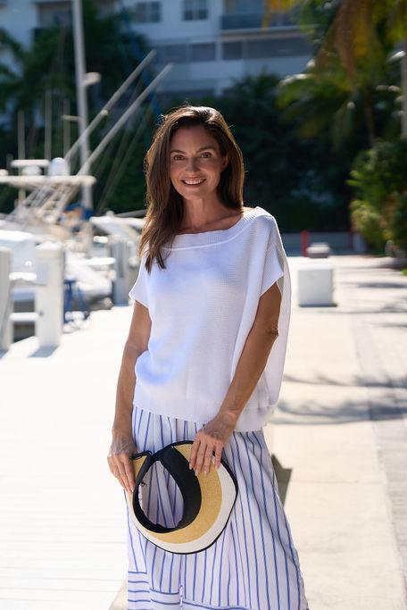 Woman in a white pima cotton sweater holding a hat by a marina with boats and palm trees in the background in Fort Lauderdale, Florida.