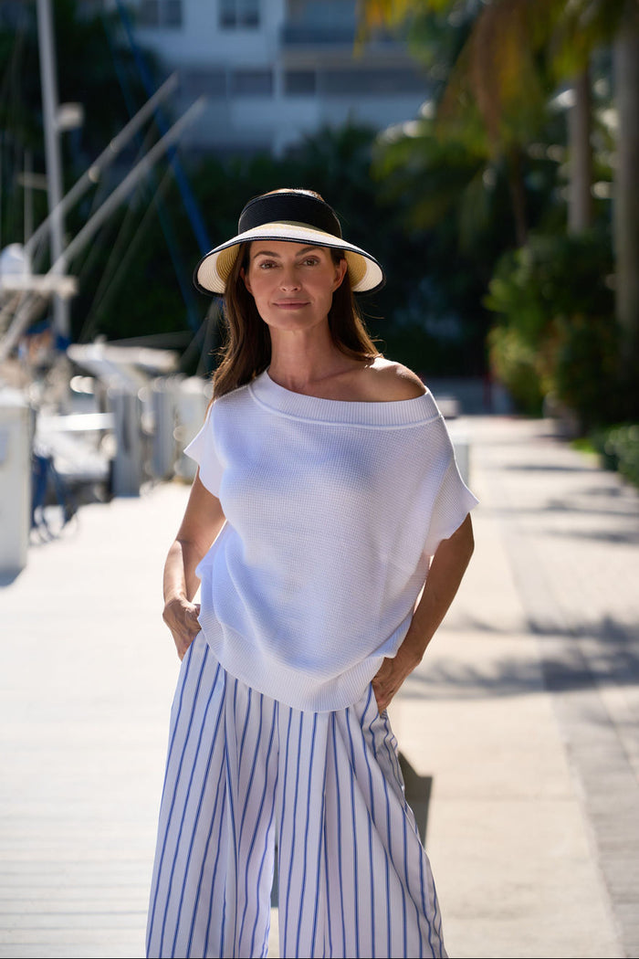 Woman wearing a white pima cotton knit top and striped pants with a black and white hat, standing on a sunlit path in a Fort Lauderdale marina.