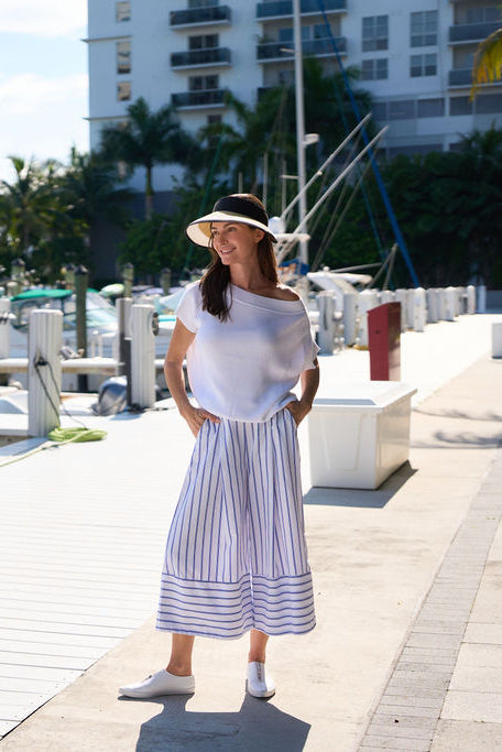 Woman in a white pima cotton knit top and striped pants standing on a dock with buildings and palm trees in the background.