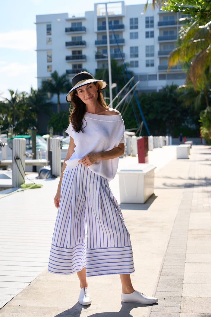 Woman in a white off-shoulder Pima cotton top and striped pants standing on a waterfront promenade.